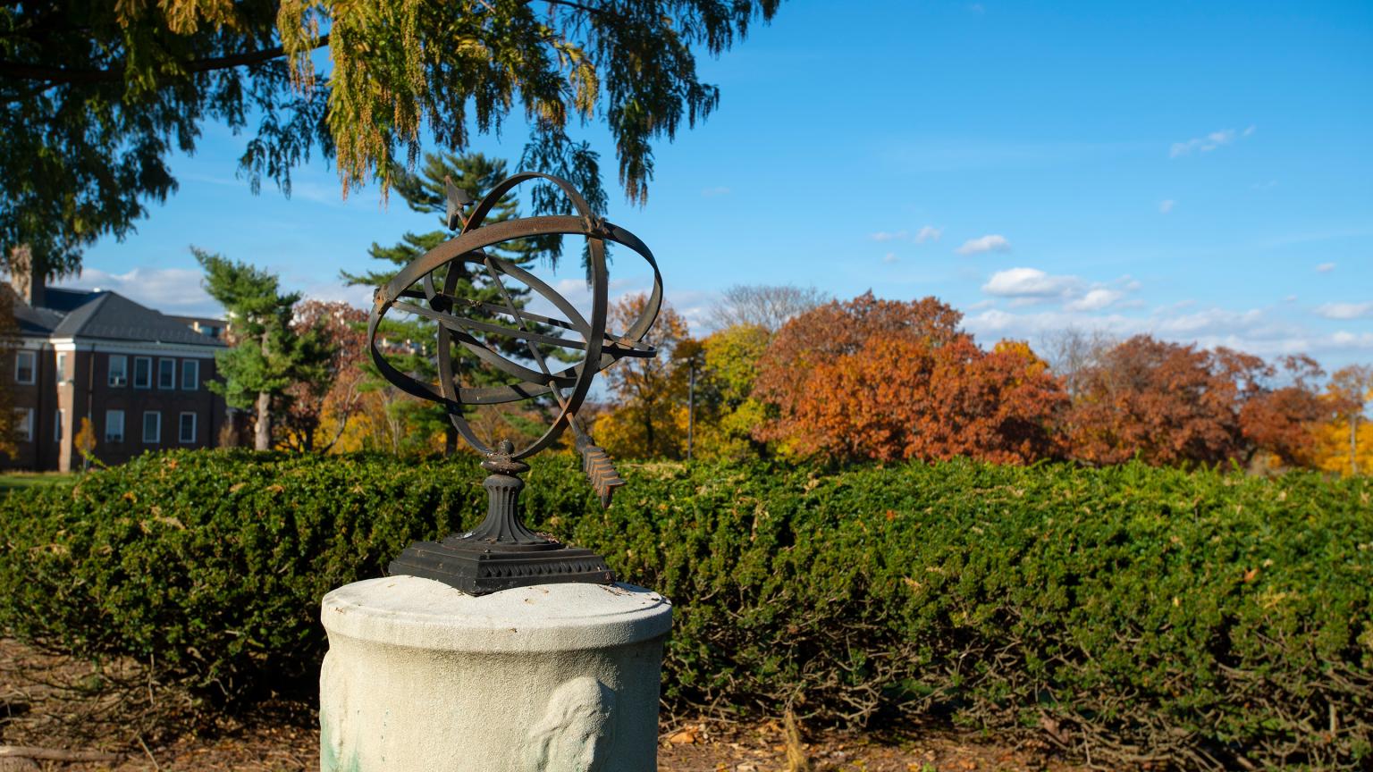 Sundial sculpture against fall trees under a bright blue sky on the Douglass Campus