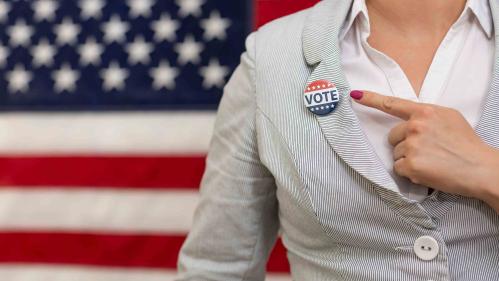 A woman standing in front of an American flag points to a "Vote" button on her lapel.