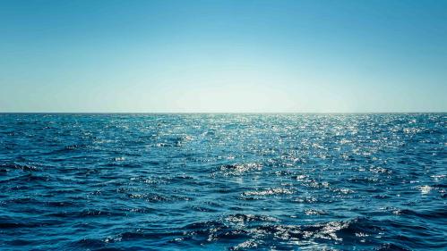 A wide expanse of blue ocean with a medium blue sky with brightness at the horizon, suggesting a sunrise.