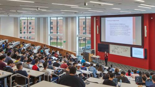 A classroom with stadium seating filled with students with a professor at center teaching