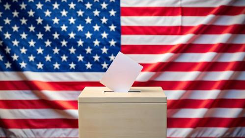 A ballot falls into a ballot box in front of a United States flag.