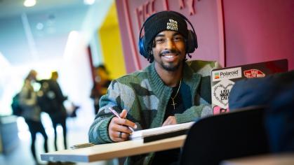 A student wearing headphones sits at a table in the Academic Building and studies on a laptop.