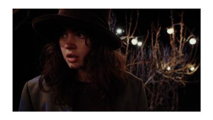 Person with long hair and hat outside in dark with lighted tree in background