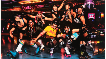 A group of indigenous women post for a photo in a roller rink. They are all dressed in 80s attire, high socks, helmets, bright colored t shirts and black tank tops. They all wear roll skates.