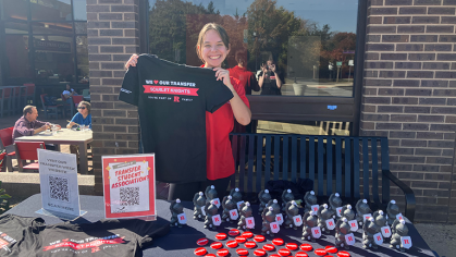 A student sitting behind a table with water bottles and pins, holding up a t-shirt that says "We love our Transfer Scarlet Knights. You're part of R family"