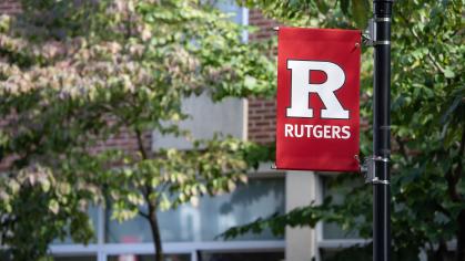 A red banner with the Rutgers block R logo hangs on a lamp post on campus against a background of green trees
