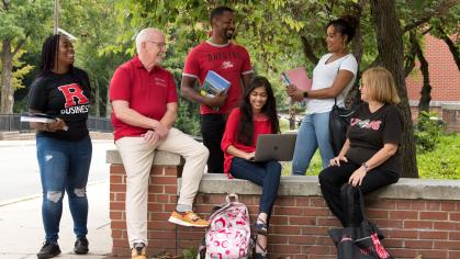 A diverse group of individuals wearing Rutgers apparel sitting on and standing around a short brick wall