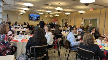 People seated at round tables in a well-lit conference room with folders and drinks on tables; banner and flag in background.
