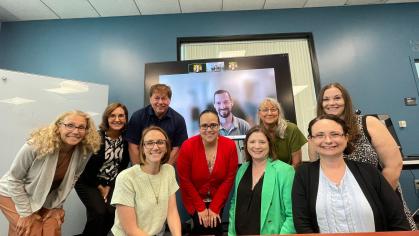 Nine people in a professional setting gathered around a conference table and one on a large screen, with blue walls and a whiteboard in the background.