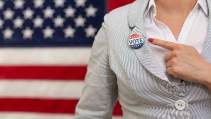 A woman standing in front of an American flag points to a "Vote" button on her lapel.