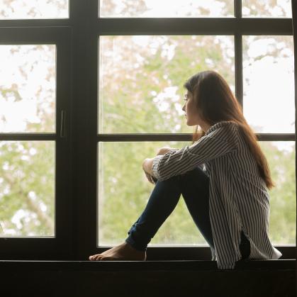 thoughtful girl sitting on sill embracing knees looking at window