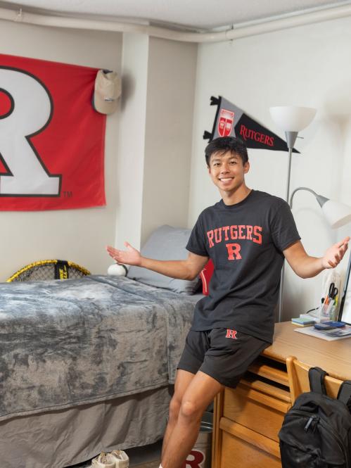 Rutgers student Ethan Thai leans on a desk in his dorm room. He is wearing a black shirt with Rutgers on it, and is surrounded by Rutgers gear
