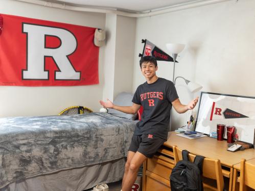 Rutgers student Ethan Thai leans on a desk in his dorm room. He is wearing a black shirt with Rutgers on it, and is surrounded by Rutgers gear