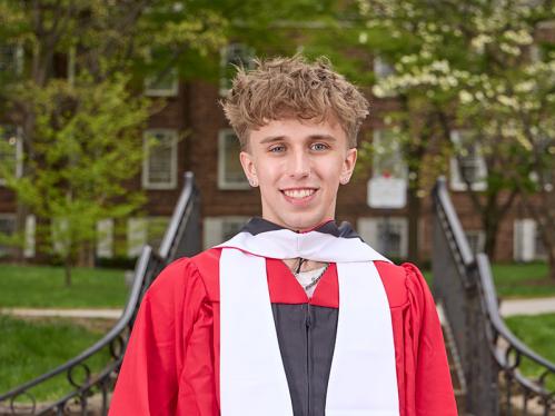 Tyler Nordquist in front of the Old Queens gate holding his graduation cap.