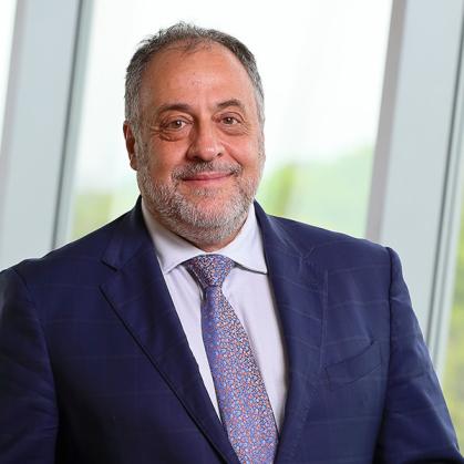 Man in a dark blue suit, white shirt and light blue tie stands in front of a window.