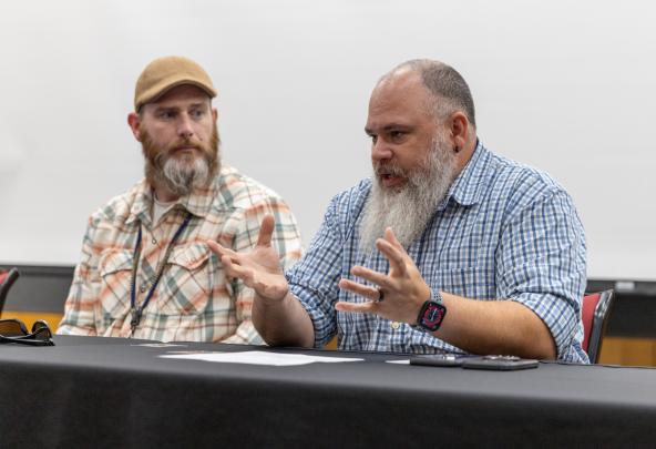 Two men with beards sit at a table during a panel discussion on service dogs for veterans. One of them is talking and the other one is listening while looking on.