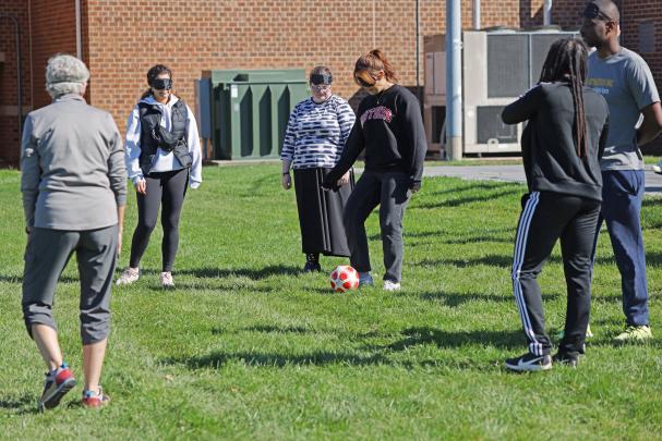 A group of people wearing eye covers kick a ball on an athletic field while playing goalball, an adaptive sport for blind and visually impaired people.