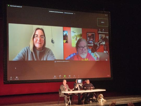 Three people sit on a stage after a screening of the film Crip Camp, while the directors are seen via video conference on a screen behind them.
