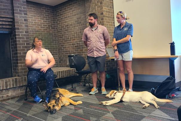 Three people share information as part of a program on guide dogs. Two guide dogs sleep on the floor next to them.