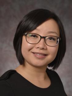 A formal portrait of smiling person with short dark hair, wearing glasses and a black top